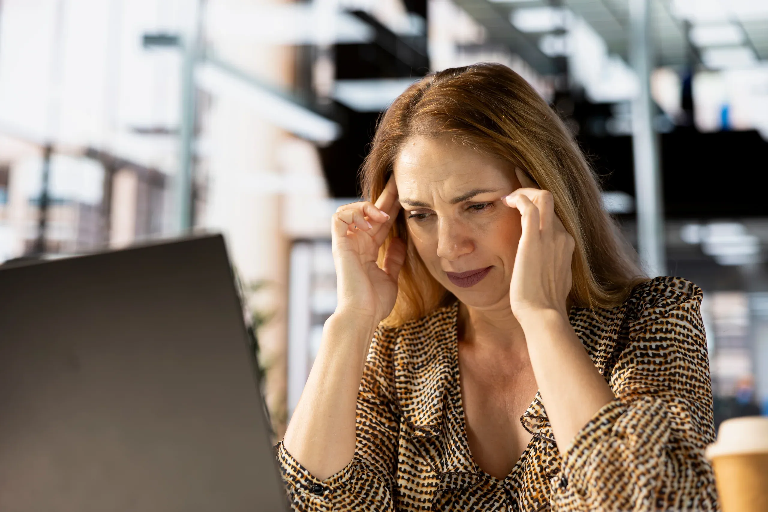 Woman at desk pressing temples while looking at laptop screen with strained expression - workplace stress and anxiety triggers