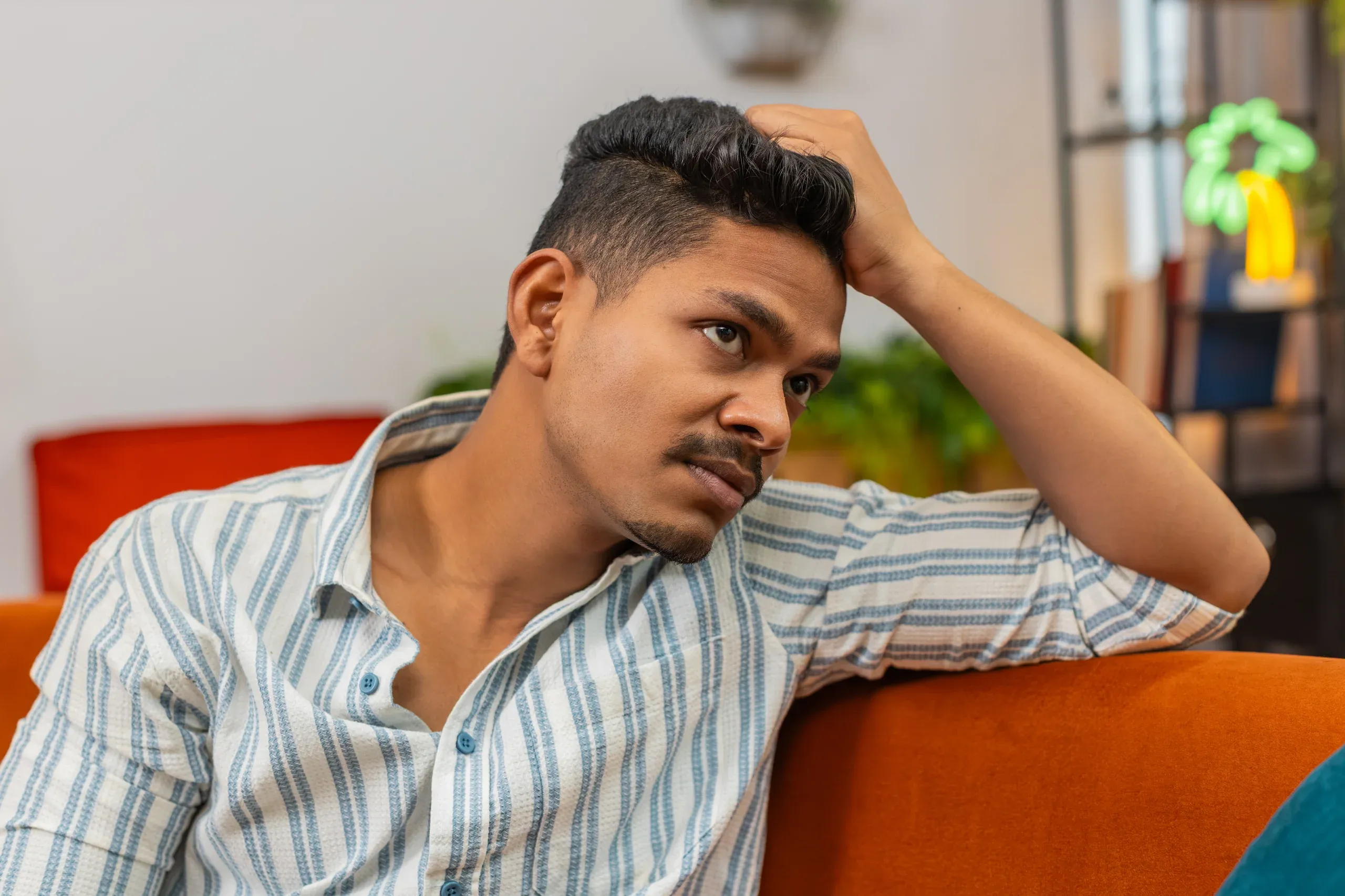 Young man on sofa with distant troubled expression running hand through hair - mental health treatment for anxiety at ANU Face Mind Body