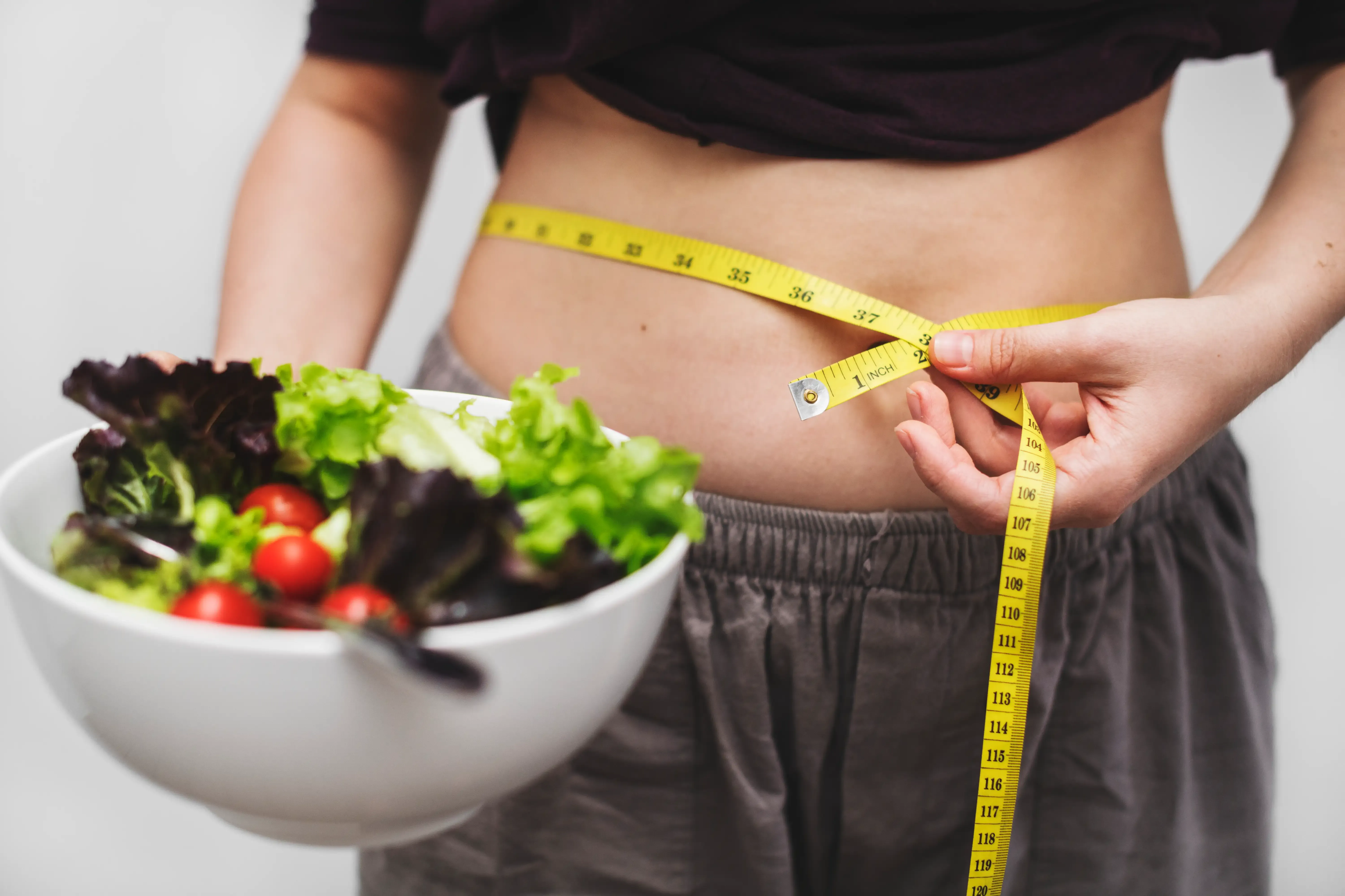 Woman holding healthy salad while measuring her waist with tape measure - metabolic health and weight loss at ANU Face Mind Body